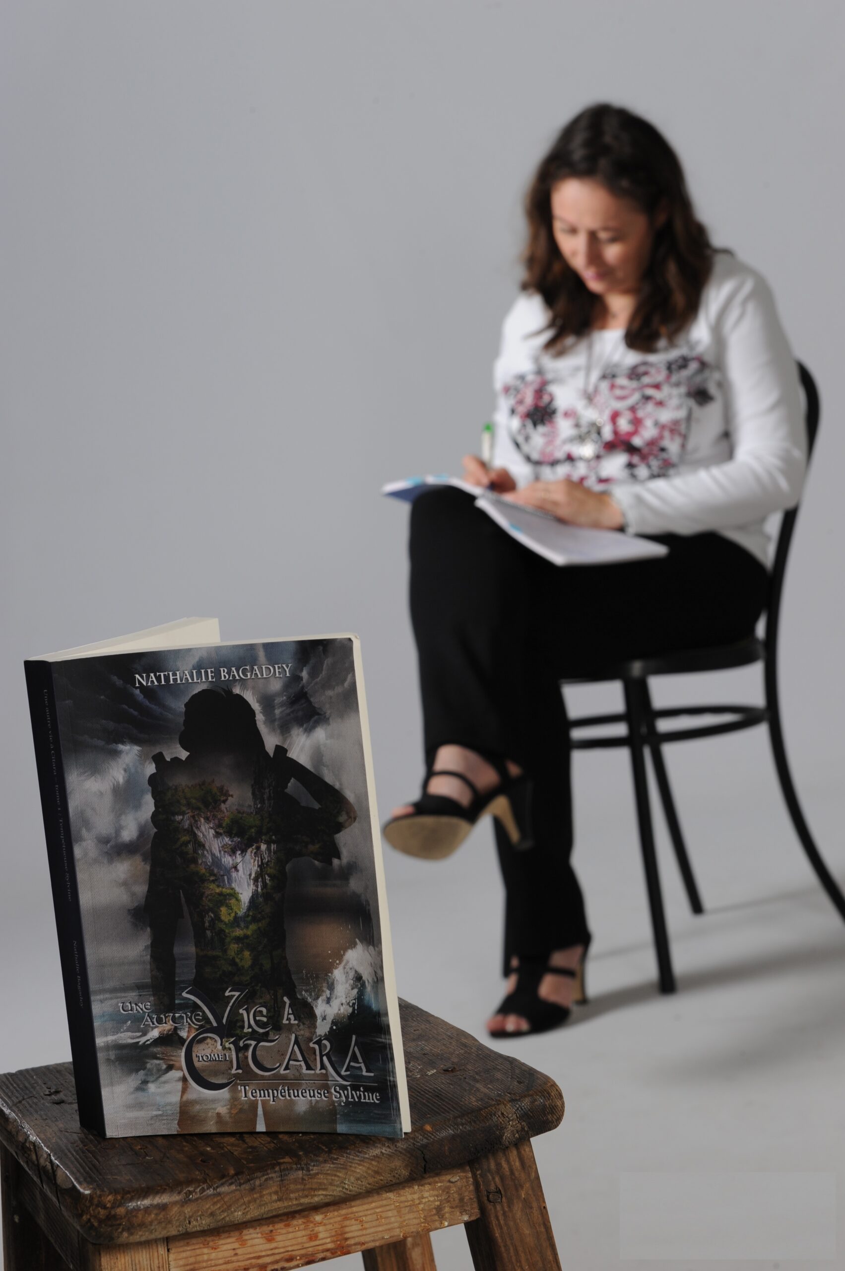 Arrière-plan : Nathalie Bagadey sur une chaise en train d'écrire. Premier plan : son livre Citara (tome 1) posé sur un tabouret.